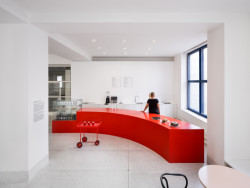 Hned vedle vstupních dveří je umístěn zaoblený sytě červený bar, který funguje jako malá kavárna. (ATLAS, No Architects)