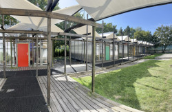 K jednotlivým učebnám se odstává po dřevěných pódiích krytých dlouhou pergolou, pro kterou byla jako „krytina“ zvolena banerovina. (Venkovní učebny GEVO na Jižním Městě, Ateliér HRA) 