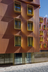 Fasáda domu kombinuje ručně taženou omítku s vertikálním žebrováním v cihlově červeném odstínu se zlatými šambránami kolem oken. (Rezidence U Milosrdných na Starém Městě od Fránek Architects)