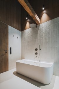 Koupelna propojuje světlý keramický obklad s tmavým dřevem a přiznanou konstrukcí krovu. (Chalupa ve stráni; Mimosa Architekti)