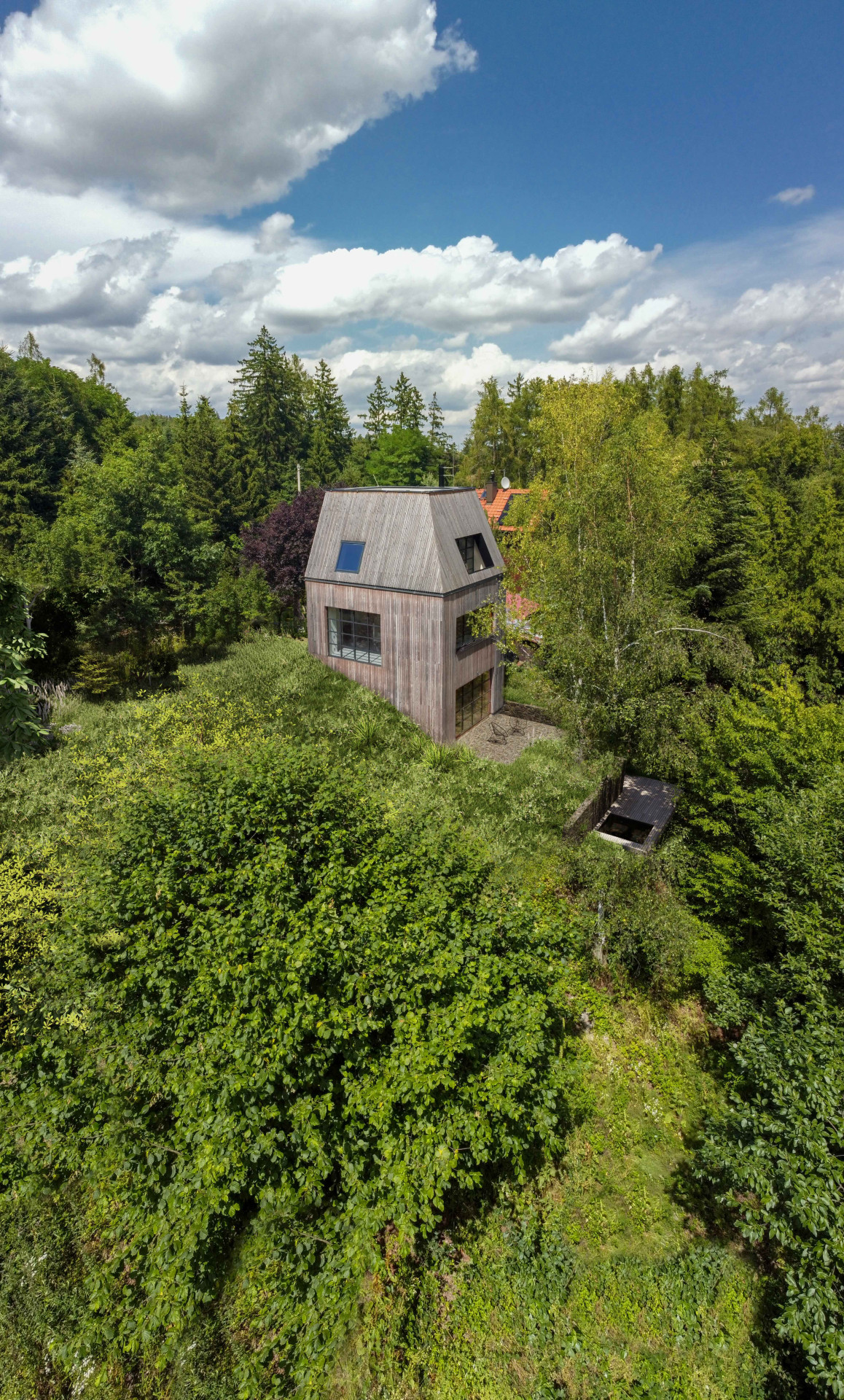 Rodinný dům je obklopený zelení. Stojí na svažitém pozemku nedaleko lesa a potoka. (Chata JARO u Prahy, edit! architects)