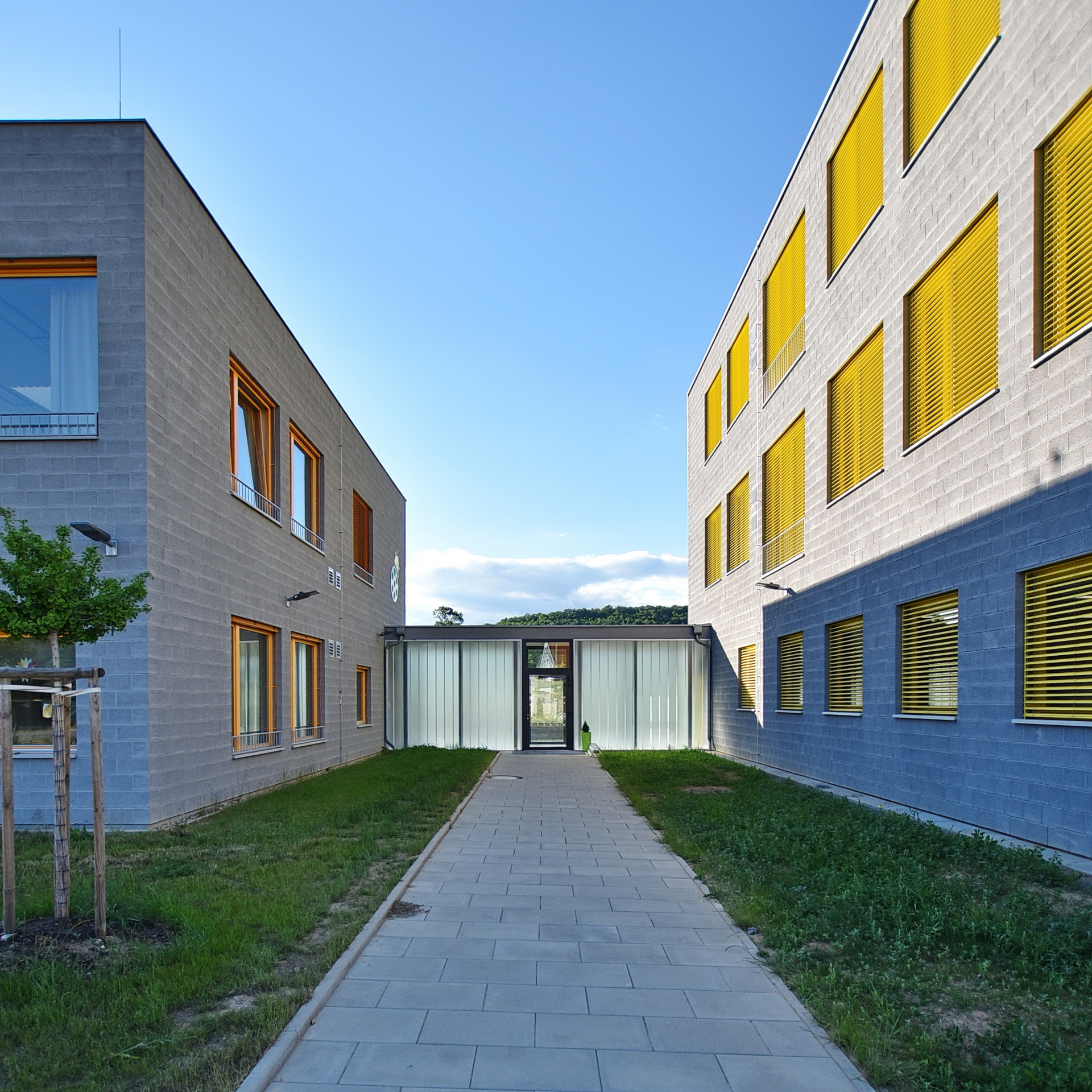 Od hravosti ještě k trošce přísnosti. Takto vypadá nová základní a mateřská škola v Letech. Šedé fasády traktují okna v pravidelném rastru, jejichž rámy a žaluzie do realizace vnášejí barvu. Škola i školka má svůj vlastní pavilon, dohromady je spojuje transparentní krček.