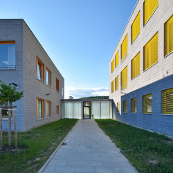 Od hravosti ještě k trošce přísnosti. Takto vypadá nová základní a mateřská škola v Letech. Šedé fasády traktují okna v pravidelném rastru, jejichž rámy a žaluzie do realizace vnášejí barvu. Škola i školka má svůj vlastní pavilon, dohromady je spojuje transparentní krček.