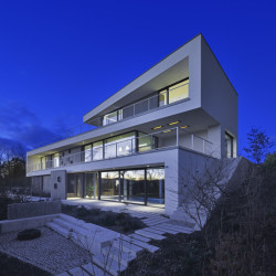 Zahradní průčelí vily se třemi podlažími a velkoformátovými proskleními, která kryjí přesahy zdiva.