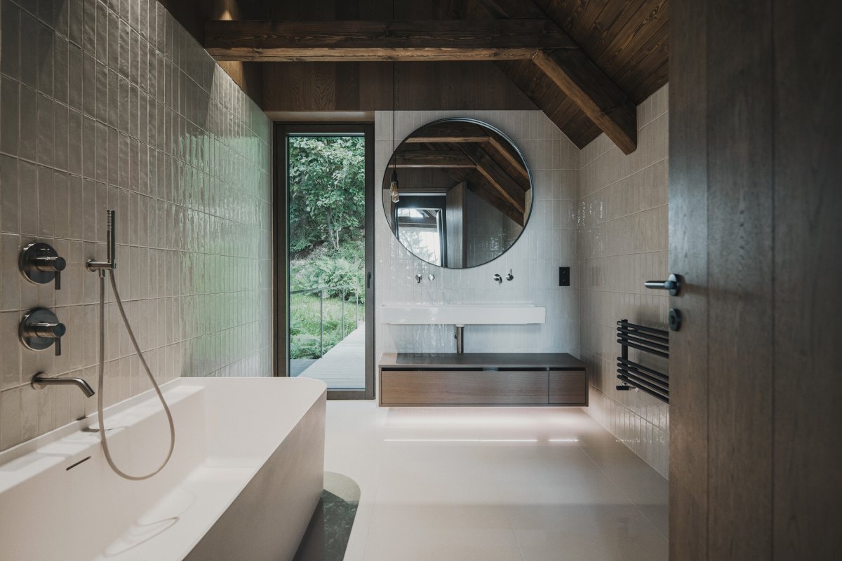 Koupelna kombinuje současné materiálové řešení s přiznaným krovem a výhledy do okolní krajiny. (Chalupa ve stráni; Mimosa Architekti)
