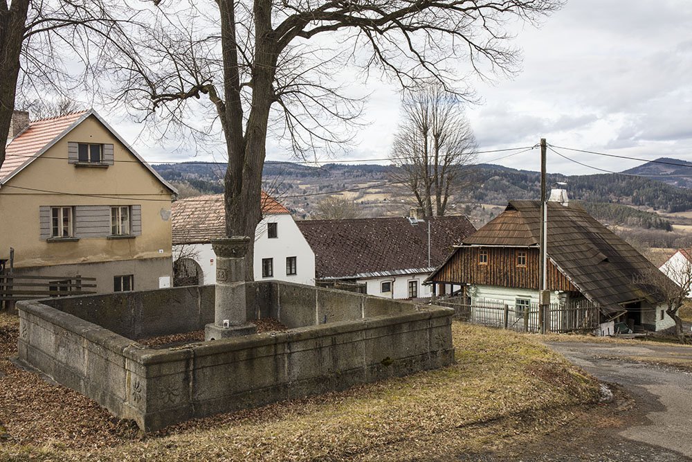 Další památkou obci Strašín je žulová kašna, roubenka v pozadí. (Roubená chalupa Strašín)