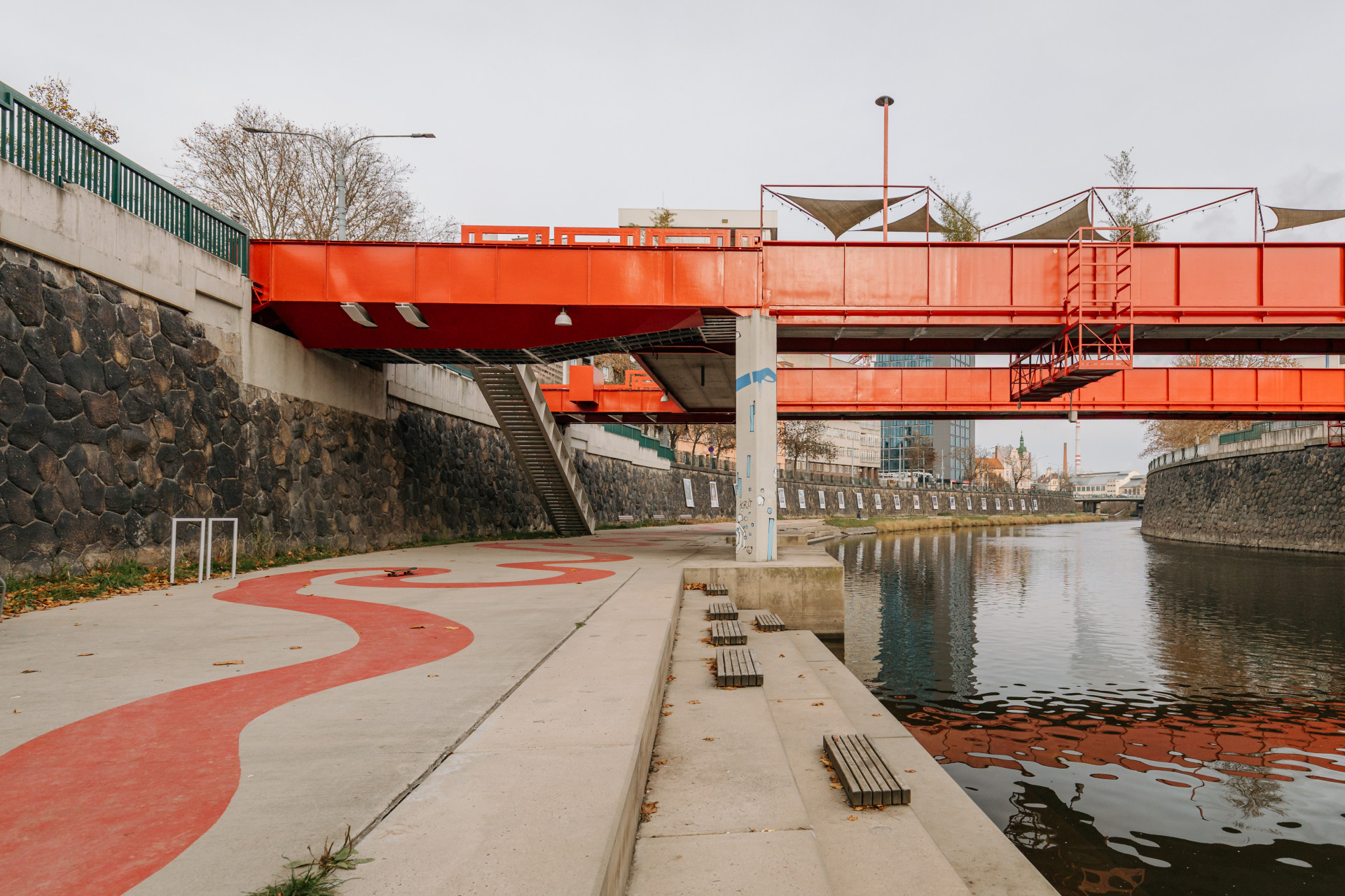Projekt zahrnuje mosty a schodiště, promenádu u řeky, plovoucí kavárnu, skate plochu i místa pro setkávání, vše sjednocené červeným motivem.