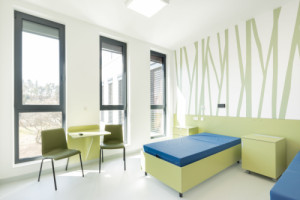 Interiéry pracují s velkými okenními plochami, přirozeným denním světlem, světlými barvami a přírodními dekory. (Pavilon psychiatrie v Ostrově od KOMA MODULAR; interiéry petit atelier)