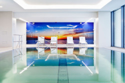 Celoskleněný obklad stěny v interiérovém bazénu. Obklad stěny o velikosti 8 × 2,5 m zahrnuje současně i troje otočné celoskleněné dveře. V momentě zavřených dveří celá stěna působí jednolitým dojmem jednoho stěnového obrazu. Umístění dveří vymezují umístěné nerezové panty a madla - foto