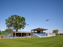 Hlavní hvězdou celé proměny je ovšem nová stavba - zázemí klubu s tribunou, která na sobě má modré sedačky, je to barva klubu. (Sportovní areál SK Planá, PROAM ARCHITEKTI)